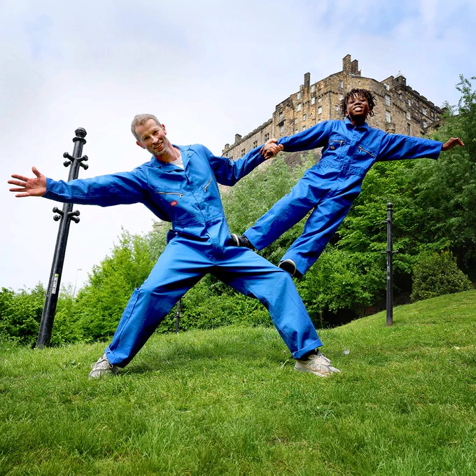 A man and a boy in matching blue overalls perform a balancing act with Edinburgh Castle in the background. The Show for Young Men, 2024. Photographer Colin Hattersley.