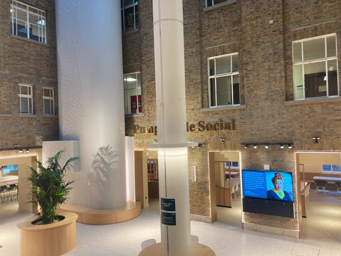 The South Courtyard area of the Pumphandle Social space. A tall plant can be seen along with seating around pillars. Part of a golden “Pumphandle Social” sign can be seen on either side of a pillar and a TV display screen can be seen too. Part of the canteen can also be seen in the background.
