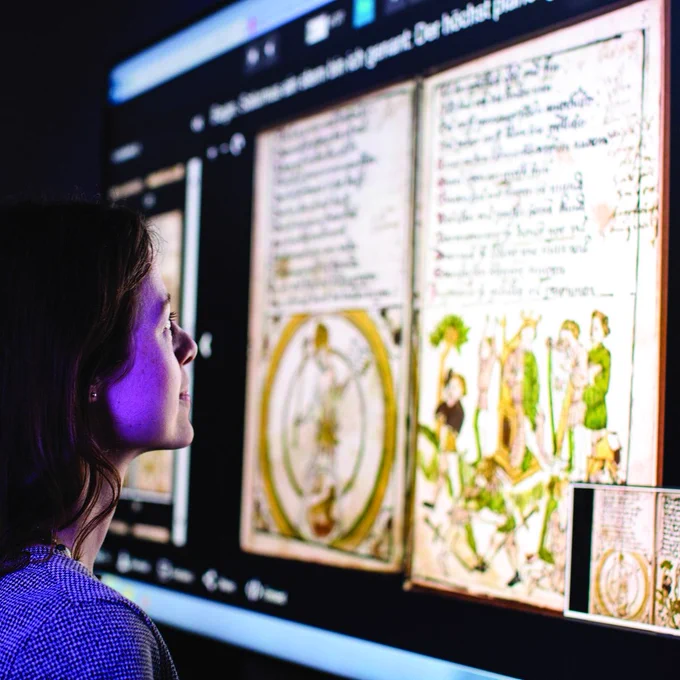 A woman looks up at a large format digital screen, which displays a digitised scan of a medieval manuscript.
