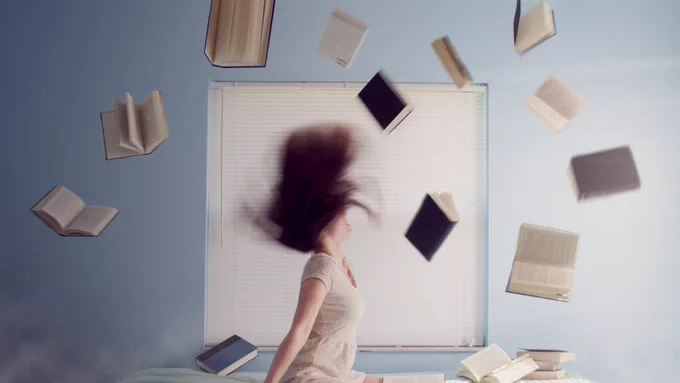 Woman sitting on bed with flying books. Photo by Lacie Slezak on Unsplash