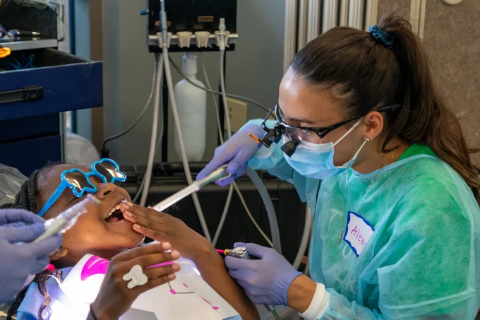 Black child smiling and laughing wearing tooth shaped sunglasses while a female dentist performs an oral exam.