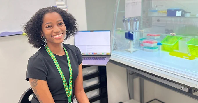 Researcher Simone Webb sits with a cheerful expression sitting at a desk in a laboratory. She is wearing a green lanyard and is in front of a laptop and lab equipment.