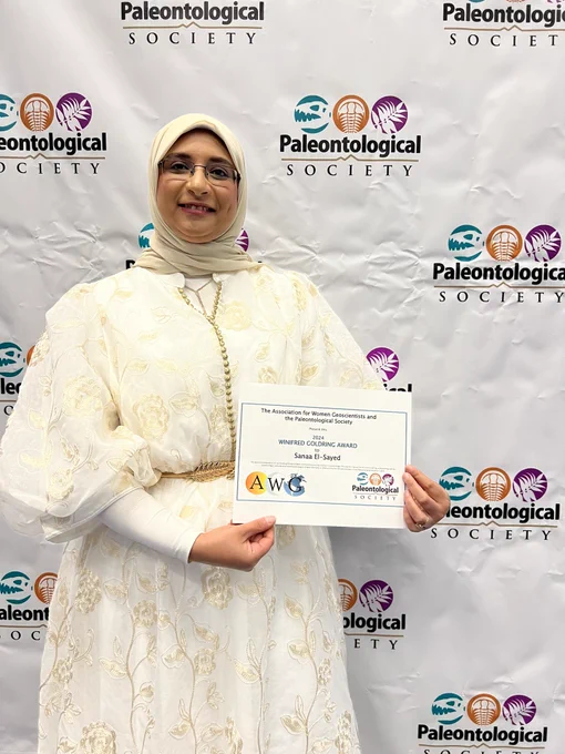 Sanaa holding her award certificate in front of PalSoc logo step-and-repeat.