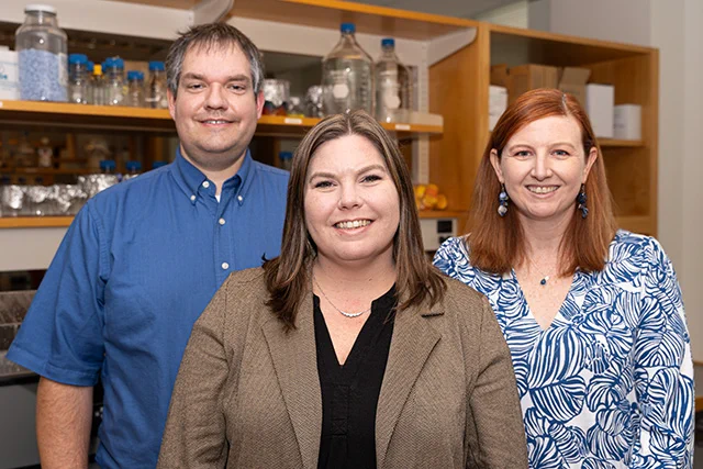 A group photo of three researchers in a lab setting. From left to right: Ben Bratton, wearing a blue shirt; Sarah Worthan, in a beige blazer over a black top; and Megan Behringer, in a blue and white patterned blouse. They are standing in front of lab equipment, smiling at the camera, showcasing their collaborative work environment.