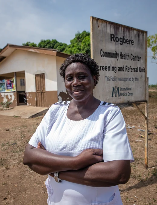 An African female nurse standing with her arms crossed outside a clinic.