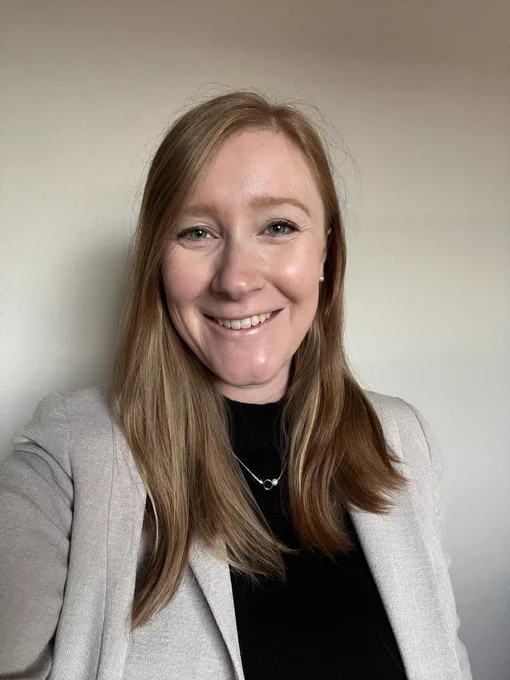 Liz in a “selfie” smiling wearing a black dress and tan blazer.