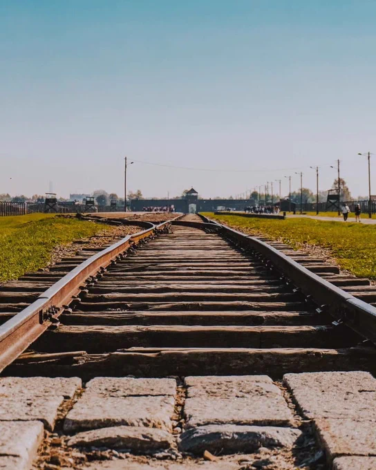 A straight railroad track stretches into the distance, surrounded by green grass fields on both sides. The sky is clear and blue, with a structure in the far distance - the main gate, creating a vanishing point. The site of the former Auschwitz II-Birkenau camp. 