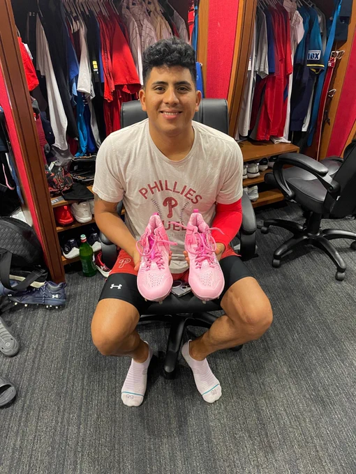 Ranger Suárez smiles as he sits in a chair in front of his locker and holds a pair of autographed cleats. He's wearing a white Phillies shirts and athletic shorts.