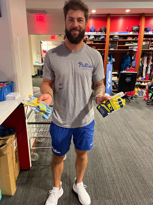 Weston Wilson smiles as he poses for a picture in the clubhouse with two pairs of autographed batting gloves. He's wearing a gray Phillies shirt and blue Phillies shorts.