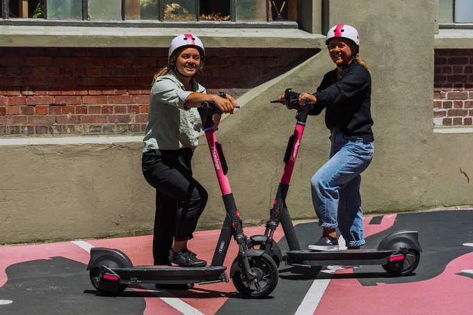 two young people smile with pink scooters