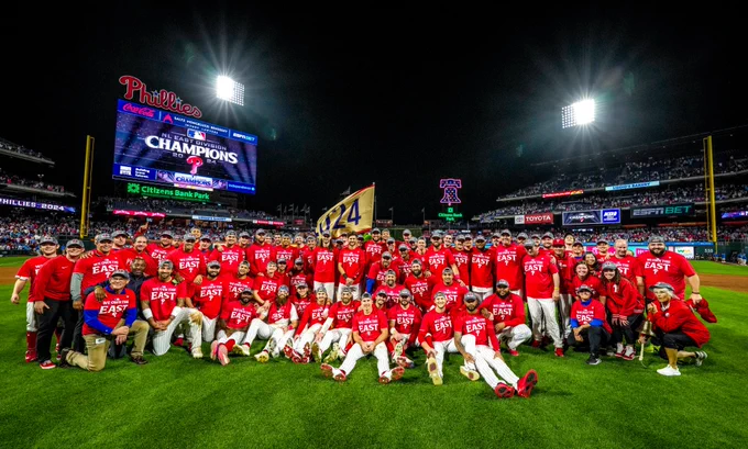 The Phillies posing together with the 2024 NL East Flag and logo on the video board in the background. Fans fill the stands to celebrate with the team.