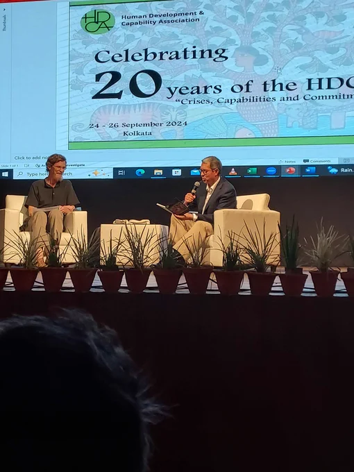 Flavio and Jean sitting on front of a screen that displays the logo of HDCA, Celebrating 20 years of the HDCA 