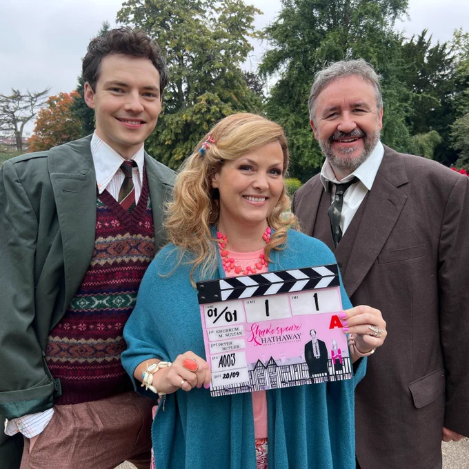 Patrick Walshe McBride, Jo Joyner and Mark Benton stand smiling for the camera in costume on set, holding a clapperboard for Shakespeare & Hathaway, illustrated with a man and a woman standing by a series of mock Tudor buildings. The trio stand outdoors in a park or gardens, smiling.