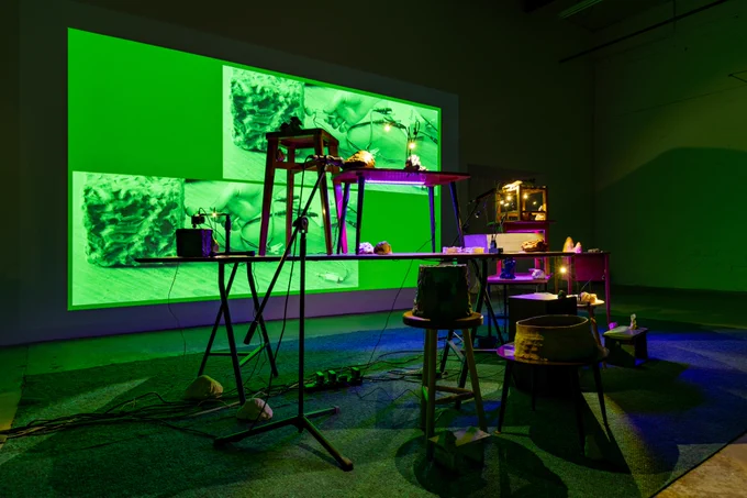 A darkened gallery with numerous objects displayed on ad hoc tables and chairs is lit by the vivid green of a wall-size video projection.