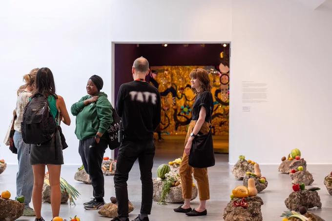 A group of people talking in a white gallery space, there are large rocks on the floor with fruit laid on top