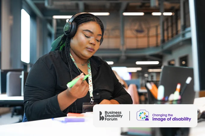 Professional wearing a sunflower lanyard to show they have a less-visible disability works at a desk using highlighters while wearing noise-cancelling headphones. The Business Disability Forum 'Changing the image of disability' logo is in the lower right corner of the image.