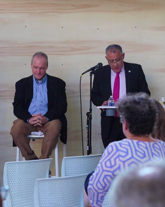 Two individuals at a public speaking event, with one seated and the other standing at a lectern, addressing an audience.