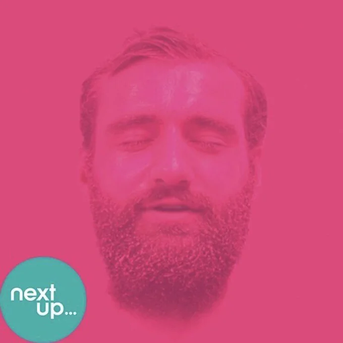 A photograph of the legendary artist Ben Target. Ben’s head is in portrait. Ben has a thick beard and Ben’s hair is in a side parting. Ben’s entire face has been painted bright pink. The background of the picture is also bright pink. This gives the impression that Ben’s head is floating. In the bottom left of the picture is a green circle with the words: Next Up in.