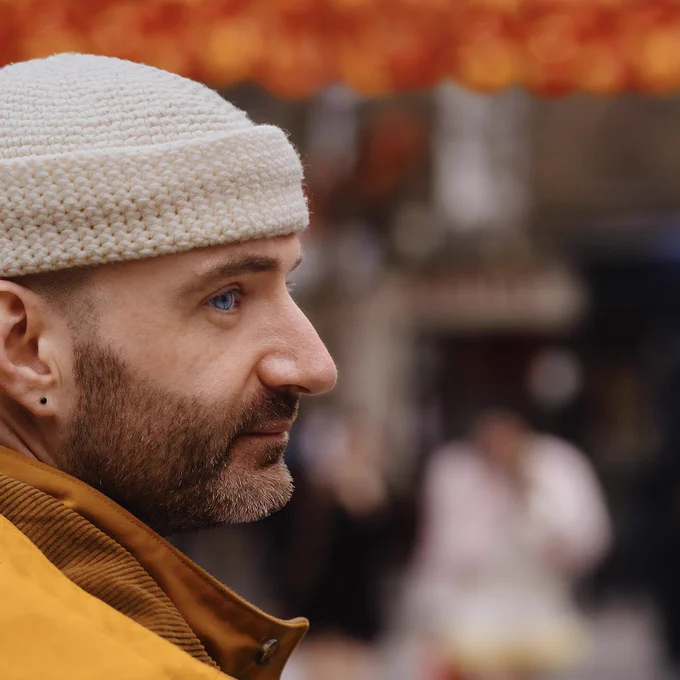 A photograph of the legendary artist Ben Target. Ben is in profile, facing from the left-hand side of the picture towards the right-hand side. In the background, there are red lanterns above Ben and two people walking towards Ben. One person is wearing a black dress, the other person is wearing a pink top and white skirt. Ben is wearing a yellow raincoat with brown corduroy lining. Ben has a short, neatly trimmed beard. Ben is wearing a pair of black, stud earrings. Ben’s eyes are bright blue. Ben is wearing a cream coloured beanie hat. Ben gazes fondly into the future.