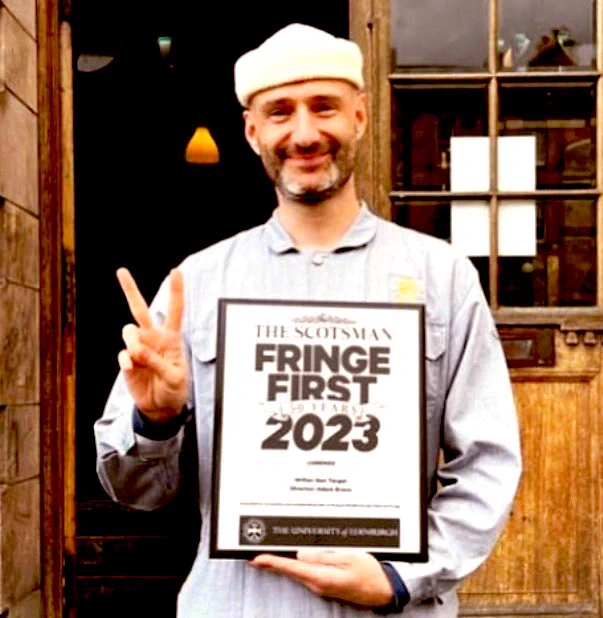 The legendary artist Ben Target holds up a placard that declares Ben the winner of a Fringe First Award for new writing for theatre. Ben makes the “peace” sign with Ben’s right hand. Ben is smiling. Ben wears a sky blue overall and a cream coloured beanie hat. Ben is standing in front of the oak doors to Summerhall in Edinburgh.