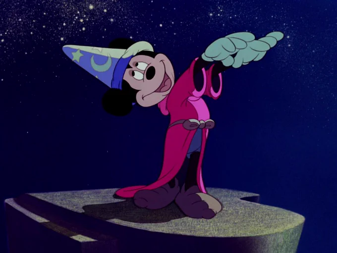 Still from Fantasia. Mickey Mouse wearing a magicians hat and a red cloak holding up his white gloved hands. He is standing on a cliff against a night time backdrop.