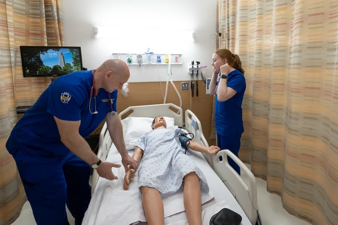 Two healthcare professionals in blue scrubs attending to a patient lying in a hospital bed, with a nature scene displayed on a television in the background.