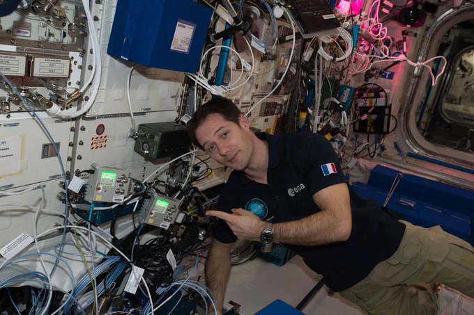 iss051e029182 (4/27/2017) --- European Space Agency (ESA) astronaut Thomas Pesquet is wearing khaki pants and a black polo shirt with an ESA emblem on the chest and a French flag on the sleeve. He is floating horizontally in the space station, facing the camera, and pointing with his left hand at the two computers, small silver cubes with green LED screens and multiple knobs on the front.
