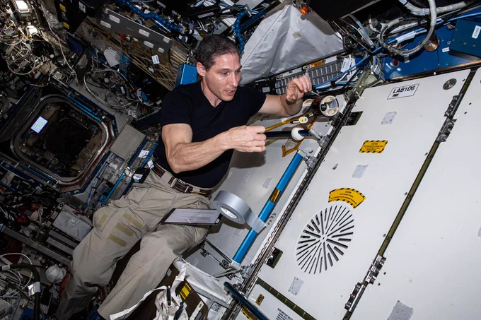  iss064e039521 (3/4/2021) --- NASA Astronaut Mike Hopkins is wearing a black t-shirt and khaki pants with a tablet attached to the thigh by Velcro. He is using his right hand to pull a strip of wide orange tape from a round dispenser attached to the wall. A roll of duct tape is stuck to a blue handrail just below his hand.