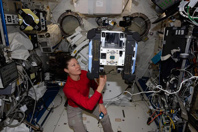 iss071e046270 (May 1, 2024) -- NASA astronaut Tracy C. Dyson, wearing a long-sleeved red shirt and khaki pants, is touching an Astrobee floating just above her left shoulder with her right hand and holding a microphone in her left hand. The robot has a blue panel on each side, black panels inside those, and a white and silver center with two camera lenses and multiple buttons on the front of it. 