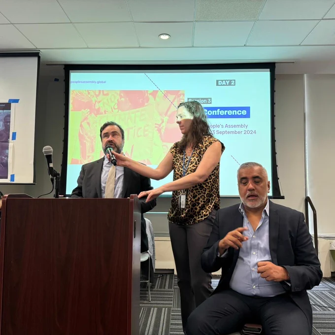 A blind Lebanese man is standing at a podium presenting in front of a room with a white woman holding the microphone and a sign language interpreter seated to the right