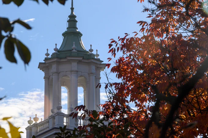 The image shows the top of a white academic building with a domed roof, rising above a tree with vibrant red leaves. The sky in the background is clear with light clouds, creating a contrast with the fall foliage in the foreground.