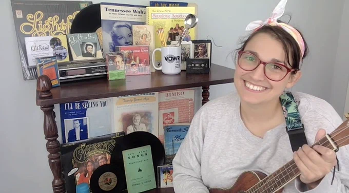 A woman posing with an instrument in front of a bookcase full of records/music/radio/tape cassettes/records
