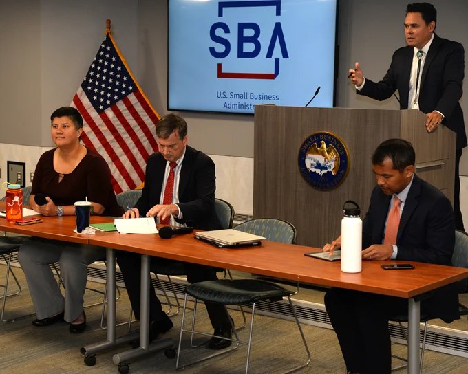 Administrator Guzman speaks at a meeting. A screen with the SBA logo and an American flag is in the background. Three others are at a table in the foreground. 