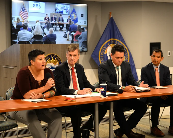 Four people sit at a table with a screen displaying the room is in the background. 