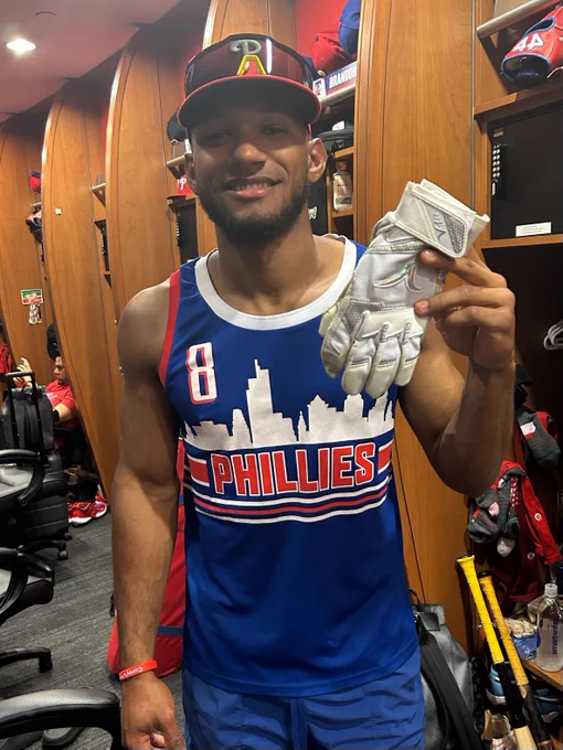 Johan Rojas smiles and poses for a picture while holding up a pair of white batting gloves in the Phillies clubhouse. He's wearing a blue Nick Castellanos basketball jersey and a Phillies hat.