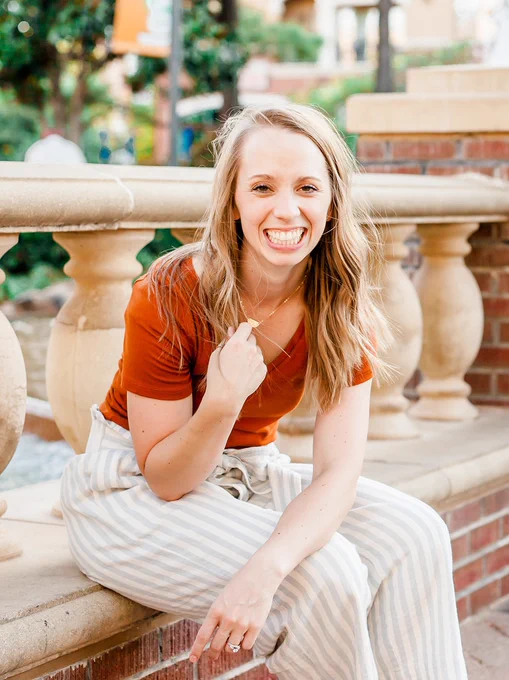 A scholar sits on a brink ledge with striped pants, orange shirt, with green trees in the background.