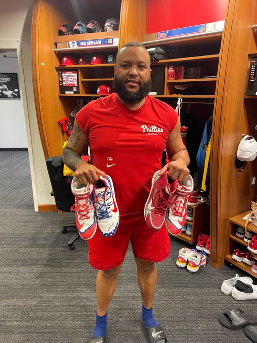 José Alvarado smiles and poses with 4 different cleats in the Phillies clubhouse. He's wearing a red Phillies shirt with matching shorts.