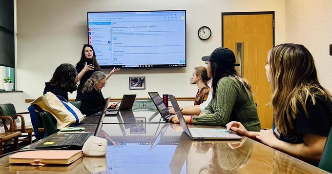 Jennifer stands in front of a screen with a KBase Narrative, talking to five workshop participants seated at a table with their laptops open. 