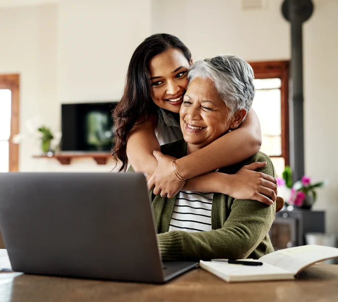 An adult daughter and her older mother share a joyful hug in front of a laptop in a home setting.