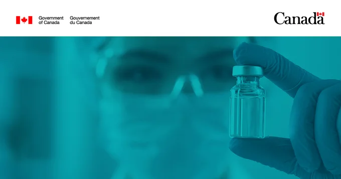 Female lab tech wearing a medical mask and safety glasses looking closely at a vaccine vial. 