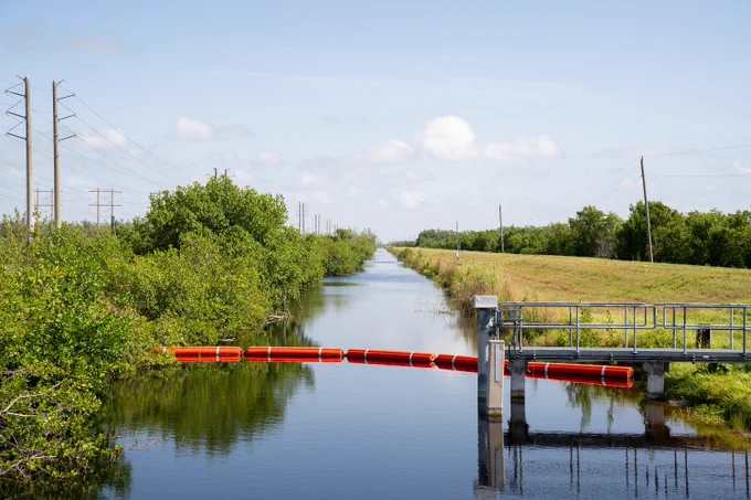 A serene landscape featuring a water canal flanked by lush greenery, with a bright red barrier across the water and a small control structure on the right. Power lines run parallel to the canal under a clear blue sky.