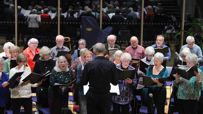 A group of choir members is shown singing together, with sheet music in hand, while facing a conductor. The conductor, seen from behind, leads them in song. The choir members are diverse in attire, some wearing bright colors and others in more subdued tones. They appear deeply engaged in their performance. The background features a reflection of the choir in large glass window.