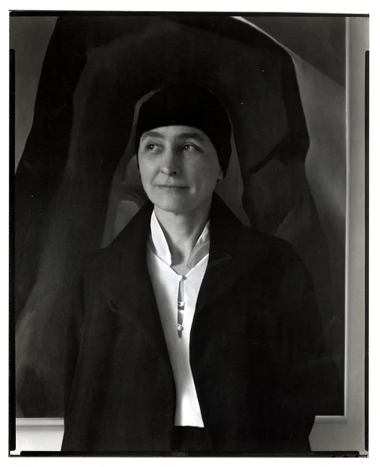 Black and white portrait of Georgia O'Keeffe standing in front of her painting, ‘After a Walk Back of Mabel's, 1929.’ The photograph was probably taken during O'Keeffe's 1930 exhibition at An American Place. O’Keeffe wears a light shirt with small buttons, a dark jacket, and a dark head covering. In the photograph, she smiles towards the left side of the frame.