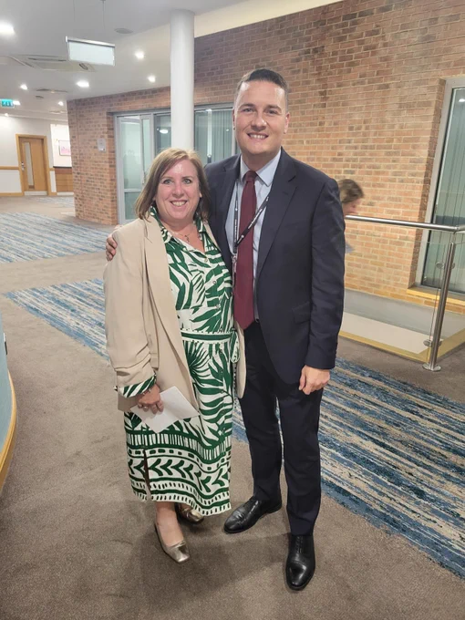 Fiona and Andrew stand side-by-side in a large hallway with exposed brick walls. Fiona has chin-length, auburn hair and wears a cream blazer over a green and white patterned dress. Andrew has his arm around Fiona. He has short, dark hair and wears a grey suit with a pale blue shirt and dark red tie. 