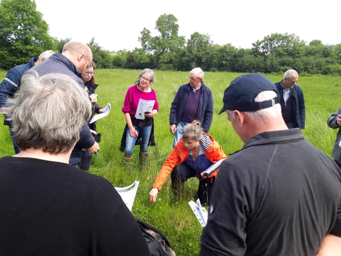 A group of people standing on a grassland surrounded by hedgerows and treelines. They are holding results-based scorecards and a person in the middle is pointing out specific indicator plants.