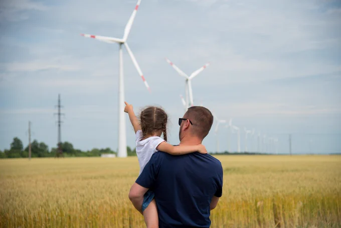 Photo of father and daughter in a field, pointing and looking at wind power and electricity pylons from distance.