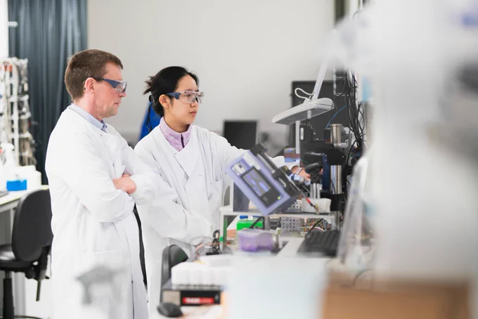 Two scientists in lab coats and protective glasses are working in a laboratory, focusing on a piece of scientific equipment. One of them is pointing to a screen while the other observes attentively.