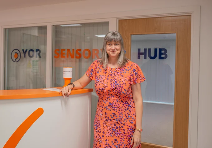 An image of a white woman, with a grey bob and straight fringe, stands next to an orange and white reception desk and rests an arm on it. Behind her is a large internal window and door with the words Yor Sensory Hub written on it.