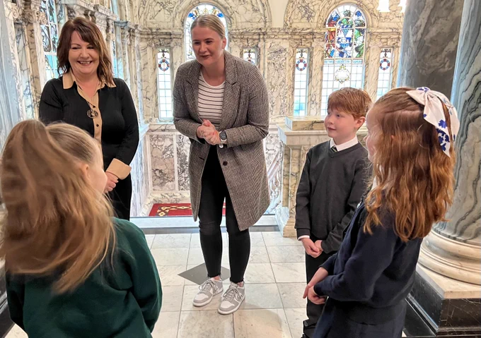 Image of five people – Junior Minister Aisling Reilly and CRIS CEO Lisa Dietrich talking to pupils from Bunscoil Bheann Mhadagáin and Cliftonville Integrated Primary School.
