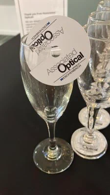 A wine glass with a disc resting on the rim with the logo for Associated Optical partner of Eschenbach. A row of wine glasses behind are resting on the same disc.